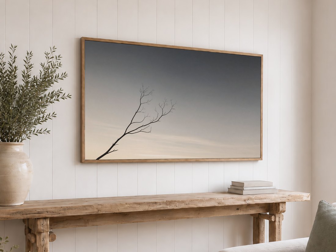 Minimalist bare branch against a soft charcoal and ivory sky shown on a light oak framed TV above a reclaimed-wood console table with olive branches in a cream vase and neutral stacked books.