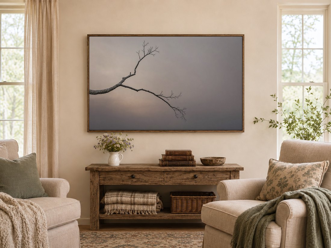 Bare branch artwork in a dark reclaimed-wood frame above a rustic console table with wildflowers, antique books and a wooden bowl, in a warm farmhouse living room with cream armchairs and sage cushions.