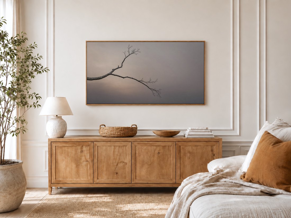 Bare branch Frame TV art in a bright bedroom with cream panelled moulding, a light oak sideboard, ceramic lamp, potted olive tree and a linen bed with a rust cushion, bringing a quiet neutral mood.