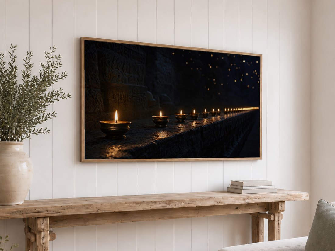Row of distant candles glowing against dark stone, displayed on a light oak framed TV over a white shiplap wall, above a rustic console with olive branches in a cream vase and stacked neutral books.