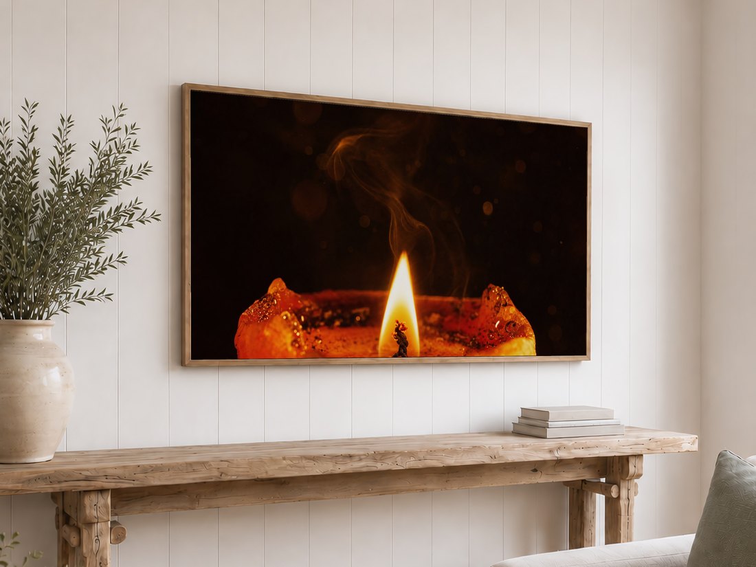 Amber candle flame close up shown on an oak framed TV against a white shiplap wall, above a rustic console table with olive branches in a cream vase and a stack of neutral books.