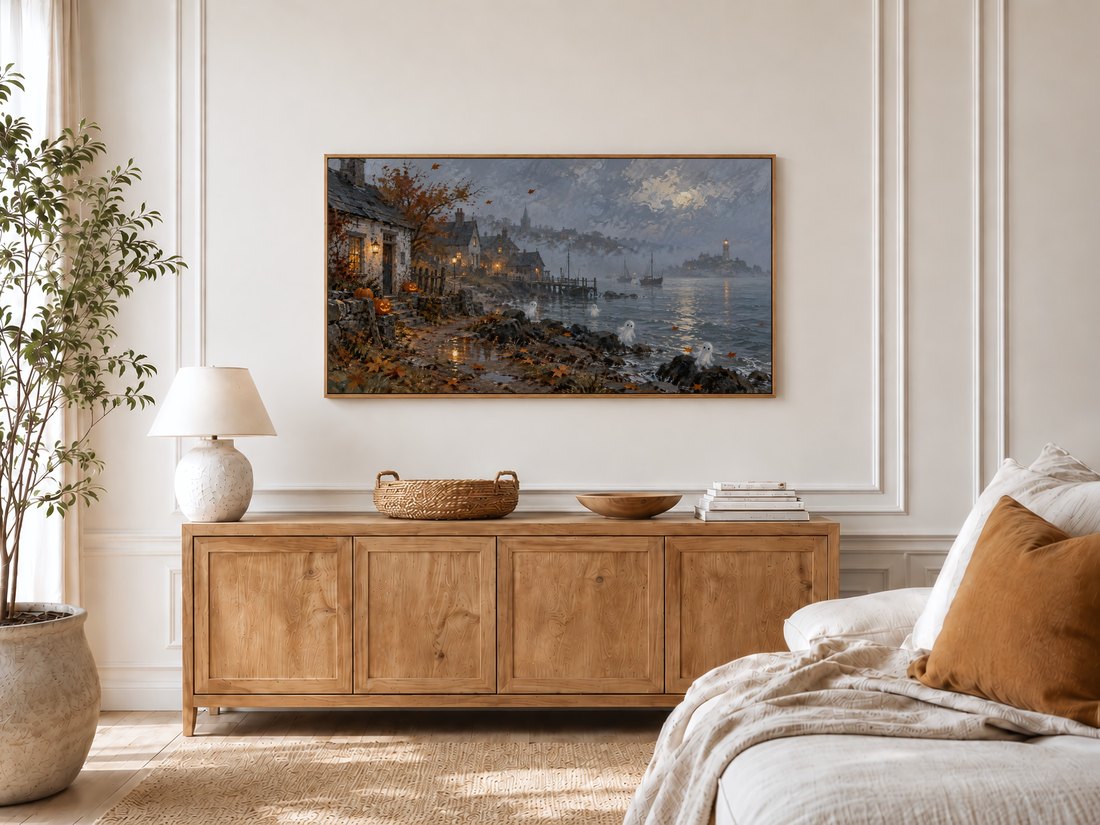 Ghostly coastal town landscape with fall foliage shown on a Frame TV in a bright bedroom with cream panelled moulding, light oak sideboard topped with textured ceramic lamp and rattan tray, potted olive tree by window and linen bed with rust cushion.