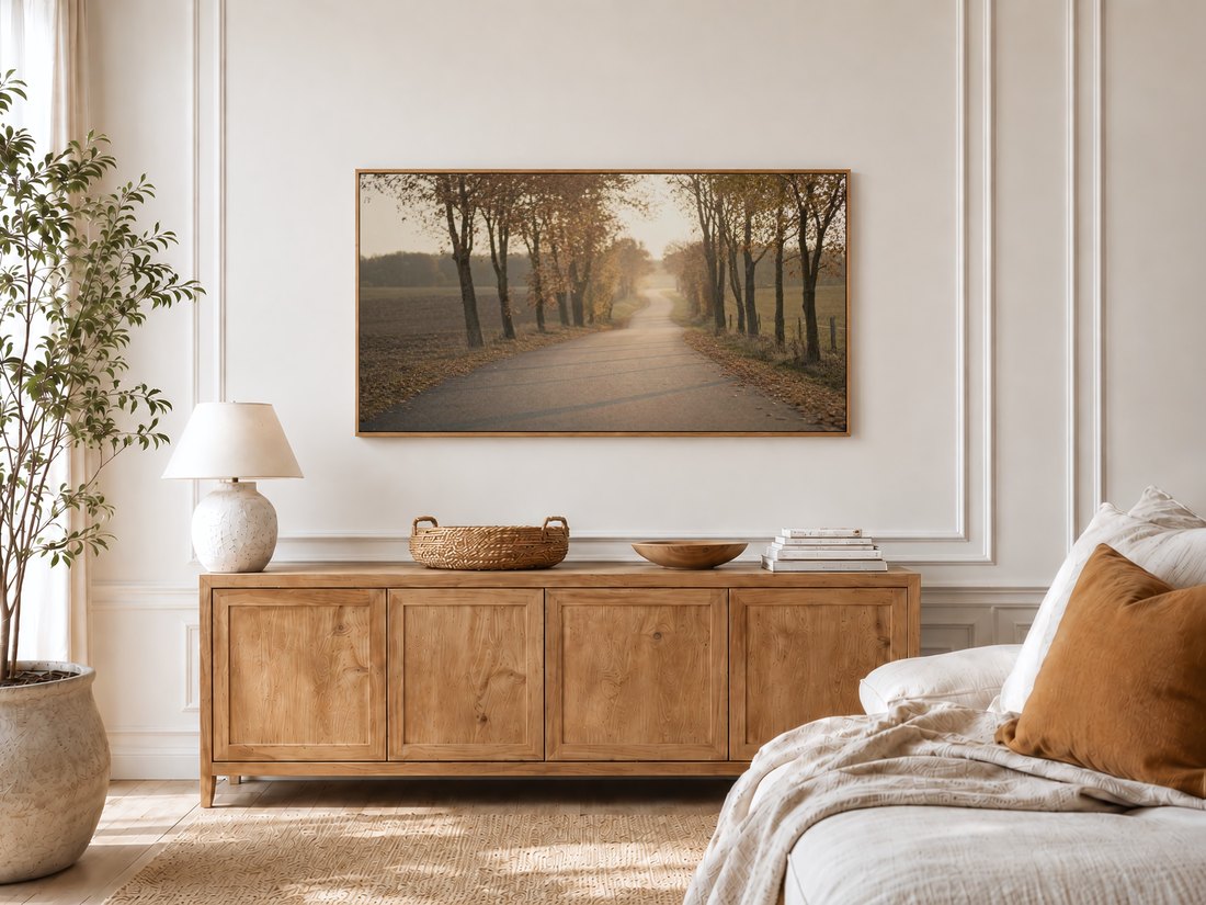 Frame TV art of a misty autumn road above a light oak sideboard with a ceramic lamp and potted olive tree, adding a calm rust and gold accent to a bright bedroom with linen bedding.