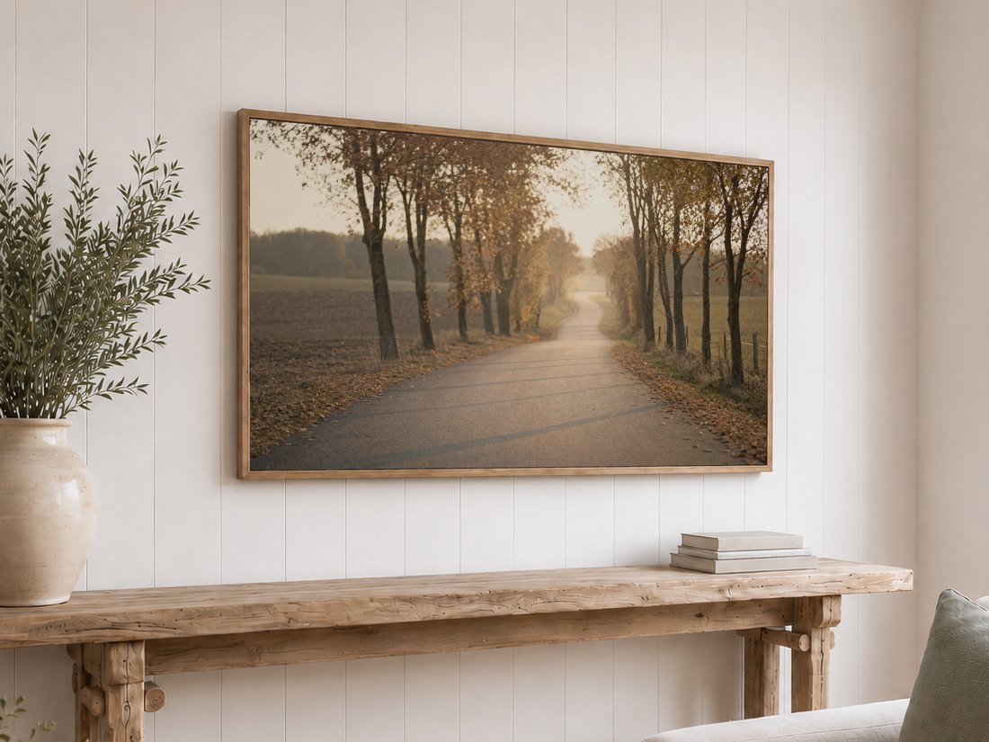 Light oak framed TV on a white shiplap wall showing a muted country road scene, paired with a rustic console table, olive branches in a ceramic vase, and stacked neutral books.