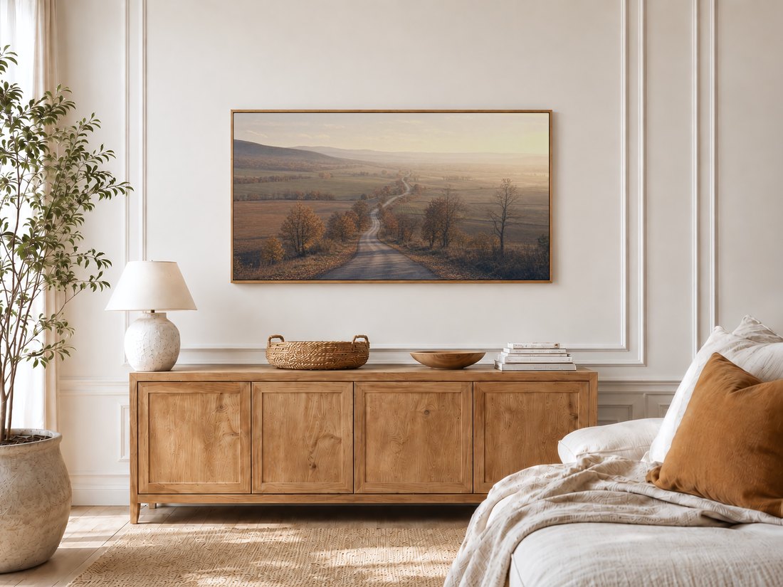 Frame TV displaying a muted autumn farmland road in a bright bedroom with cream panelled moulding, a light oak sideboard, ceramic lamp and potted olive tree, bringing a calm rustic mood to the room.