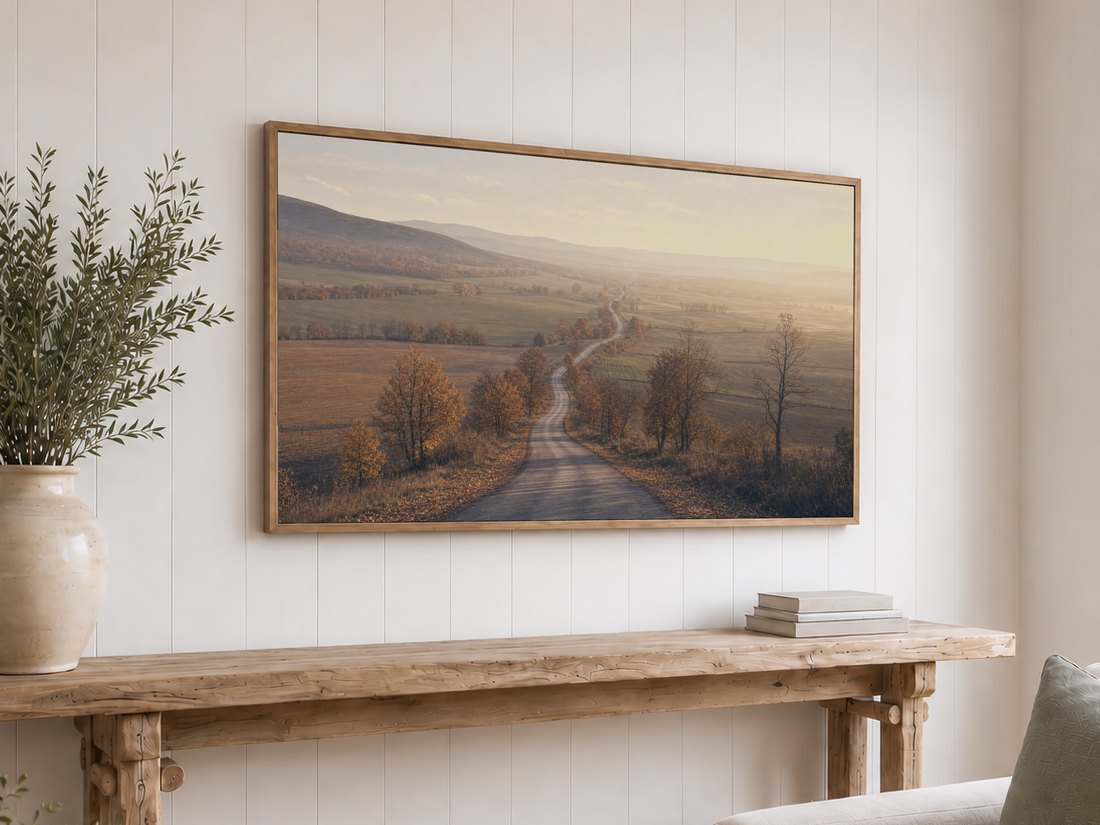 Light oak framed TV showing a soft, painterly country road scene above a reclaimed-wood console table with olive branches and neutral books, set against a white shiplap wall for a farmhouse feel.