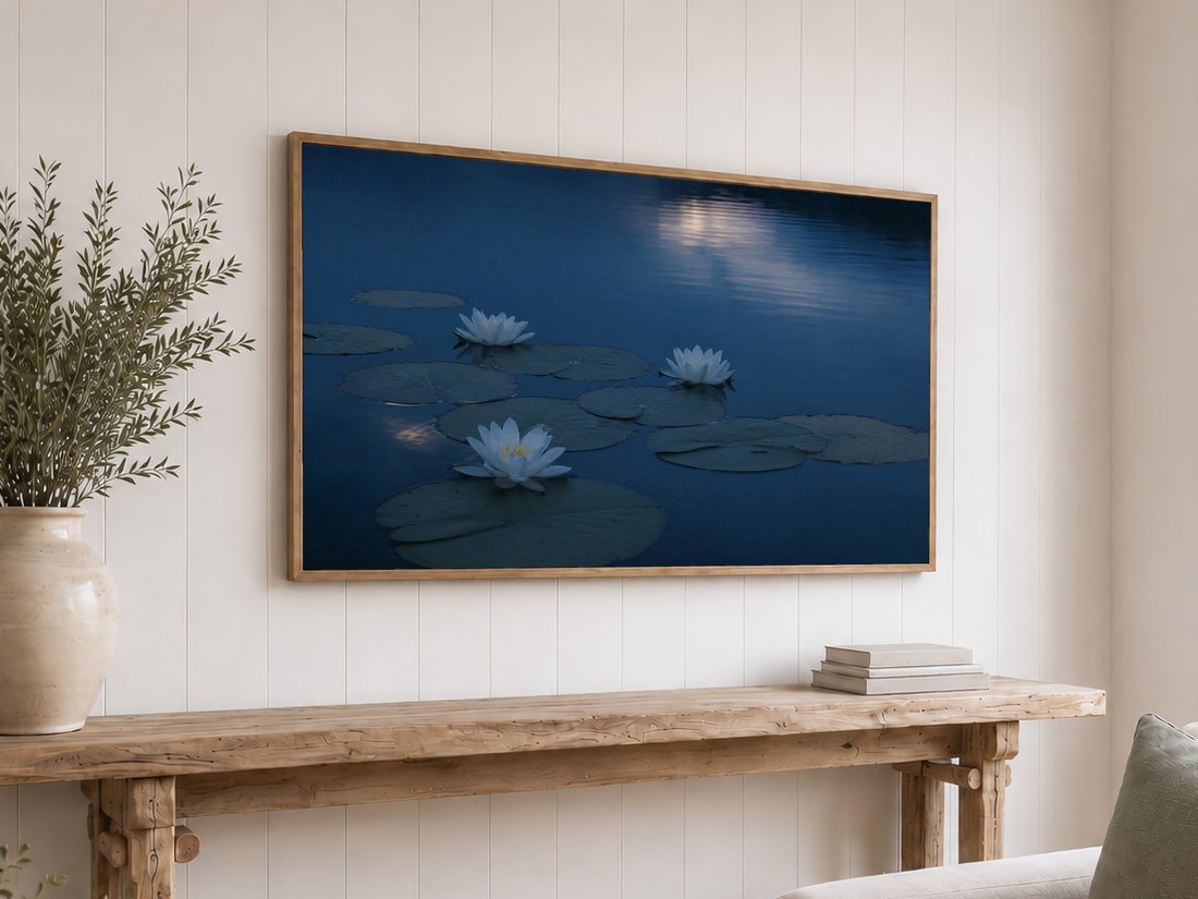 Navy and slate water lily pond art on a light oak framed TV above a rustic reclaimed-wood console table with olive branches in a cream ceramic vase and a stack of neutral books against a white shiplap wall.