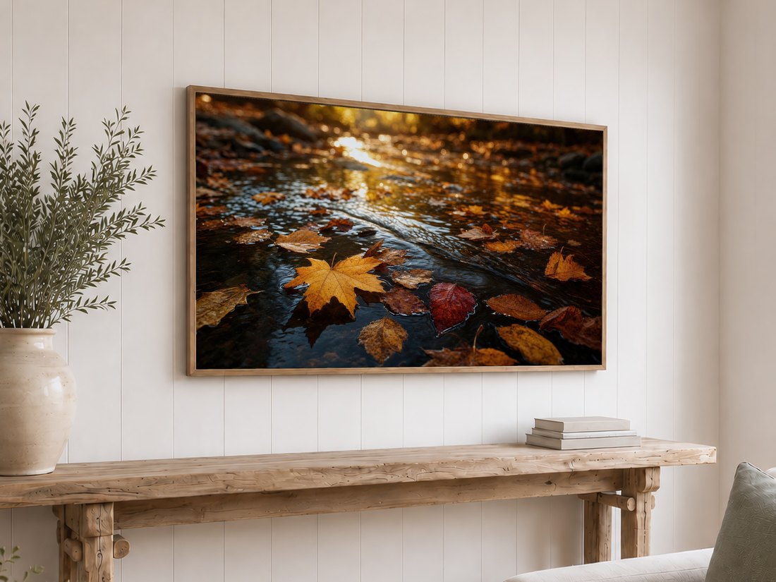 Autumn leaves on a slow stream shown on a light oak framed TV above a reclaimed-wood console table with olive branches in a cream vase and a stack of neutral books against a white shiplap wall.