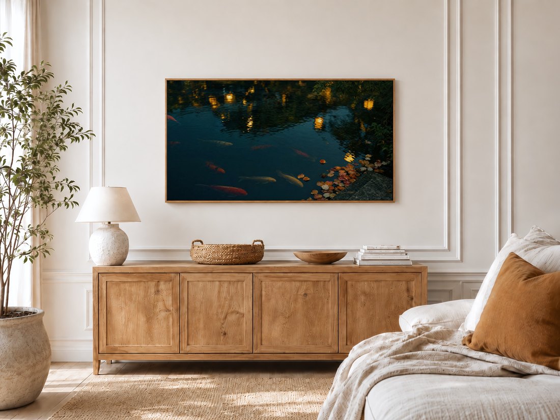 Koi pond dusk scene on a Frame TV in a bright bedroom with cream panelled moulding, an oak sideboard, textured lamp and potted olive tree, adding soft glowing color to the room.