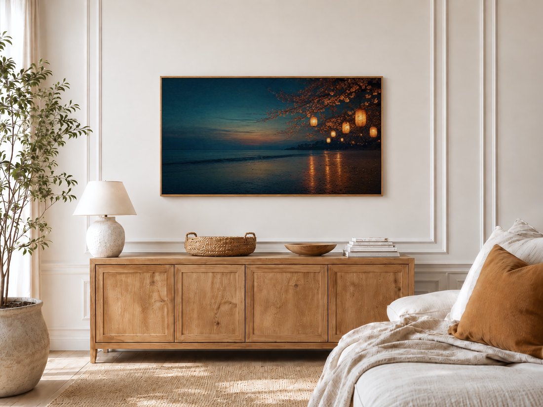 Twilight beach and blossom scene on a Frame TV in a bright bedroom with cream panelled moulding, an oak sideboard with a ceramic lamp and olive tree nearby, linen bedding with a rust cushion, bringing a quiet evening mood to the room.