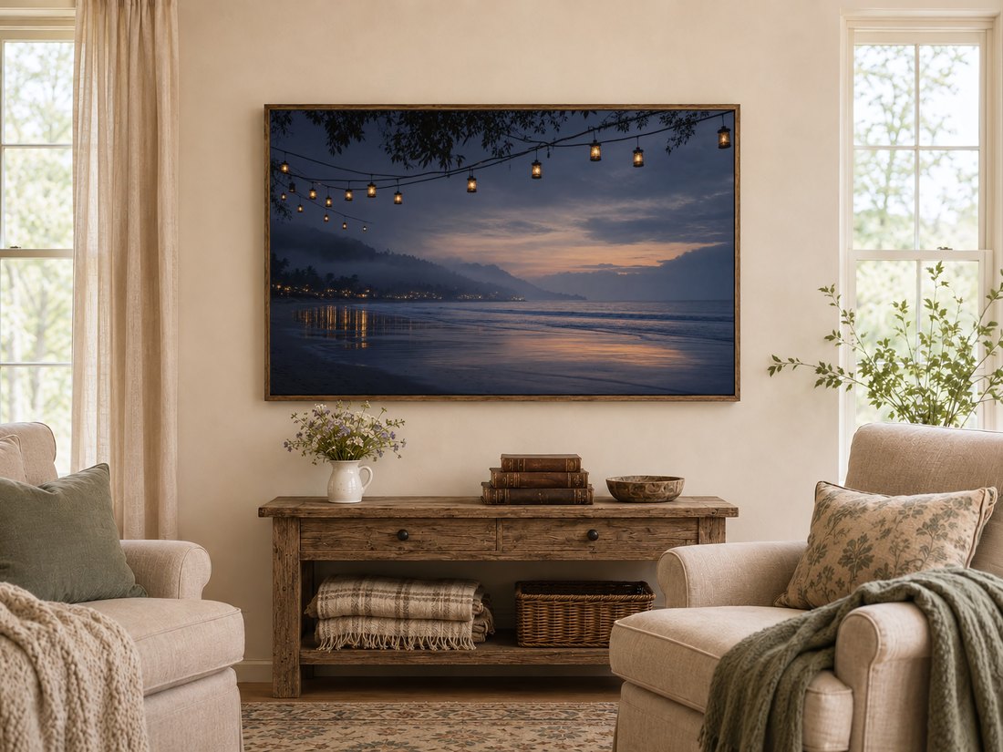 Amber-lit coastline scene shown on a TV in a warm farmhouse living room, above a rustic console with wildflowers and leather books, flanked by cream armchairs with sage cushions.