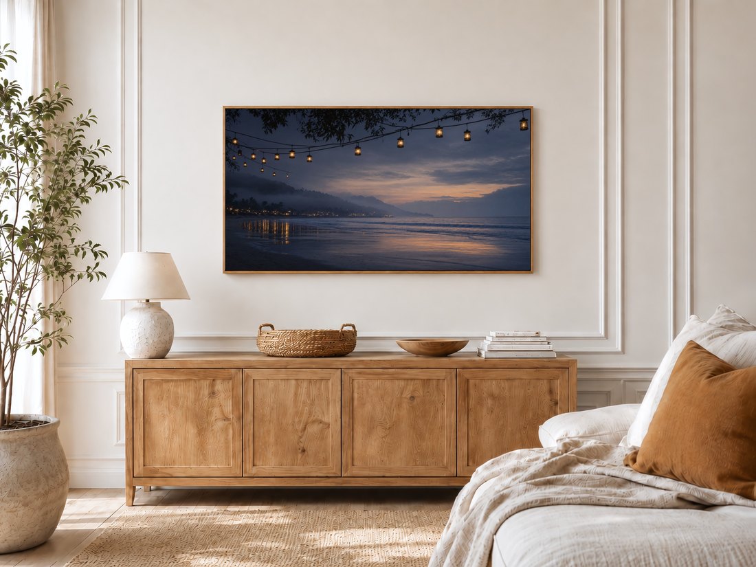 Misty lantern-lit beach art on a TV in a bright bedroom with cream panelled walls, a light oak sideboard, ceramic lamp, potted olive tree and linen bed with rust cushion.