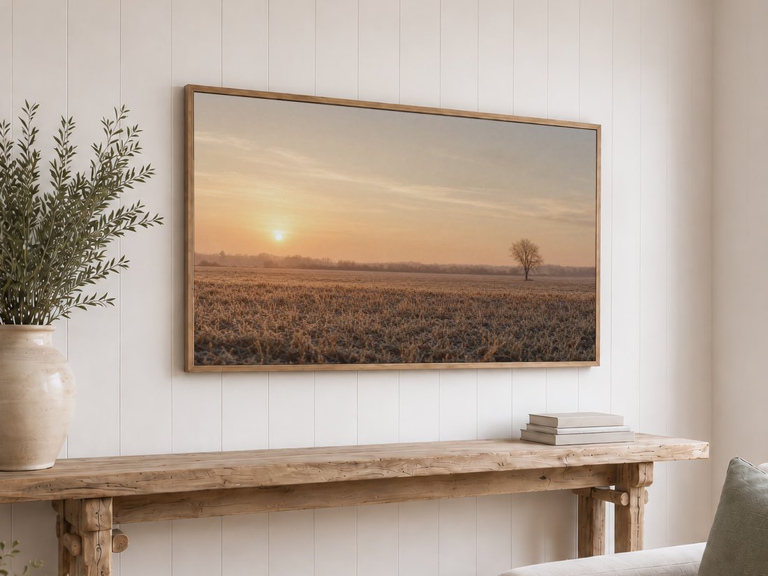 Sunrise field artwork with a distant bare tree displayed on a light oak framed TV above a rustic console table, olive branches and neutral books against a white shiplap wall.