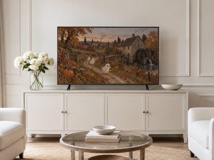 Haunted mill autumn landscape with ghosts and lanterns shown on a Frame TV mounted on a neutral cream panelled wall above a cream sideboard with brass knobs, white hydrangeas and a glass coffee table in a bright, minimalist living room