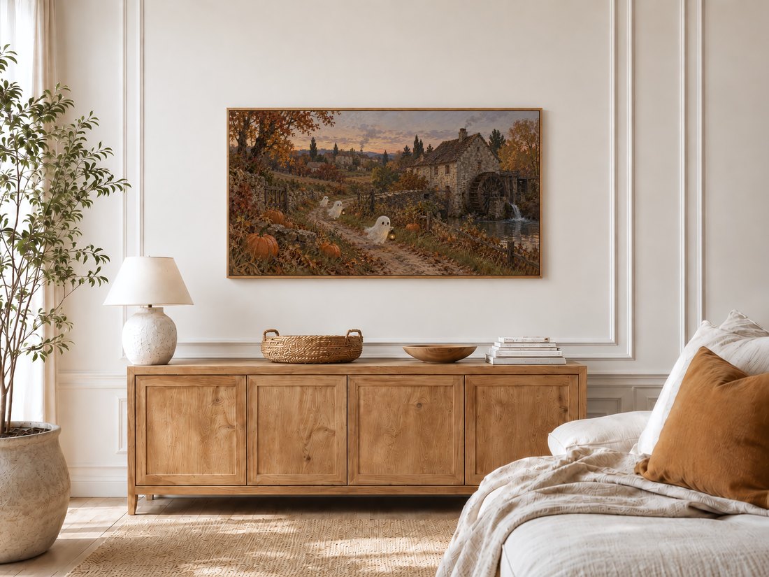 Haunted mill artwork on Frame TV in a bright bedroom with cream panelled moulding, light oak sideboard topped with ceramic lamp and wooden bowl, potted olive tree by the window and linen bed with rust cushion