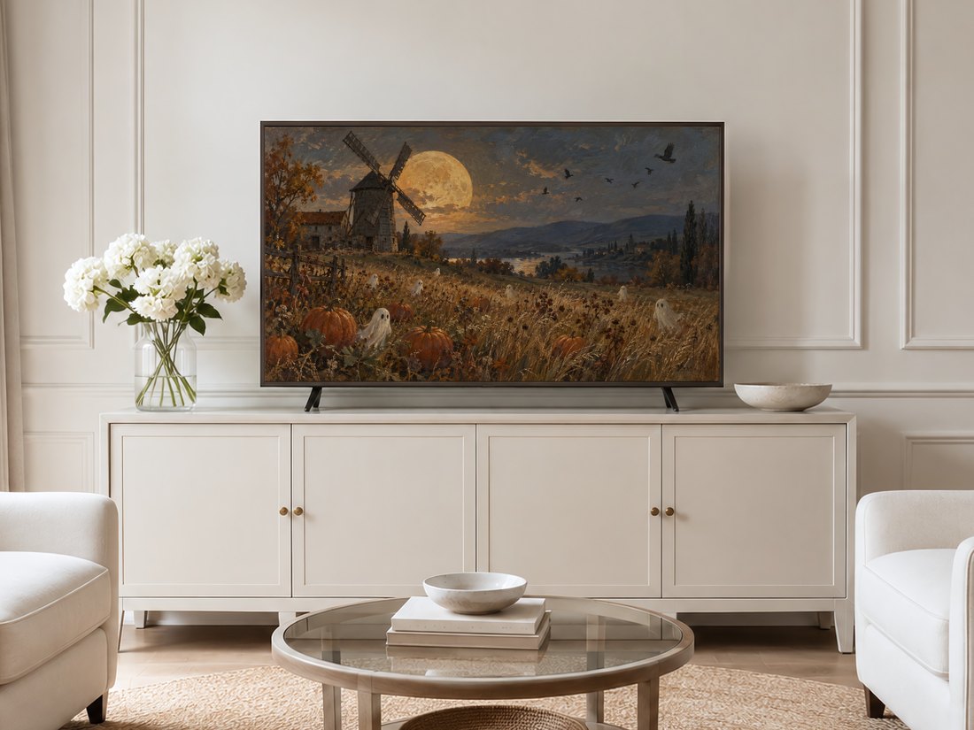 Ghostly pumpkin patch and moonlit windmill Frame TV art shown on a cream panelled wall above a light sideboard with white flowers and glassware in a modern neutral living room.