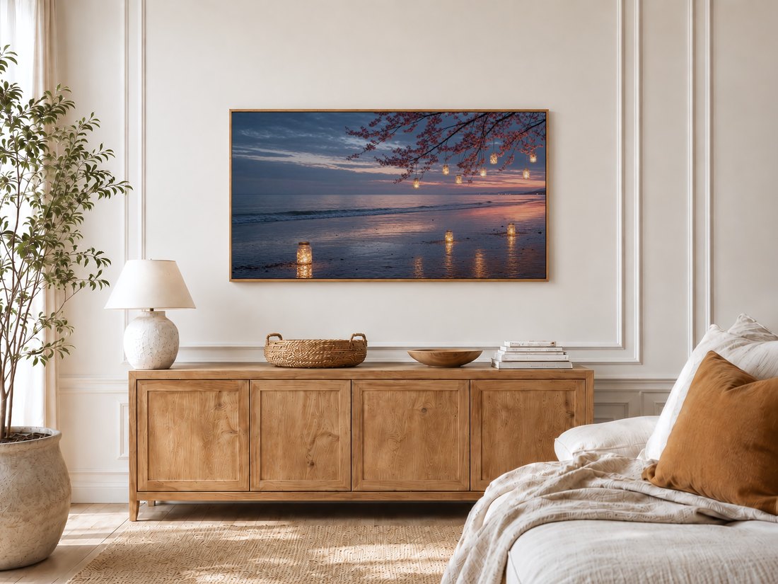 Soft coastal sunset art with lantern jars and cherry blossom branches on a Frame TV in a bright bedroom, near an oak sideboard, ceramic lamp, olive tree and linen-dressed bed.