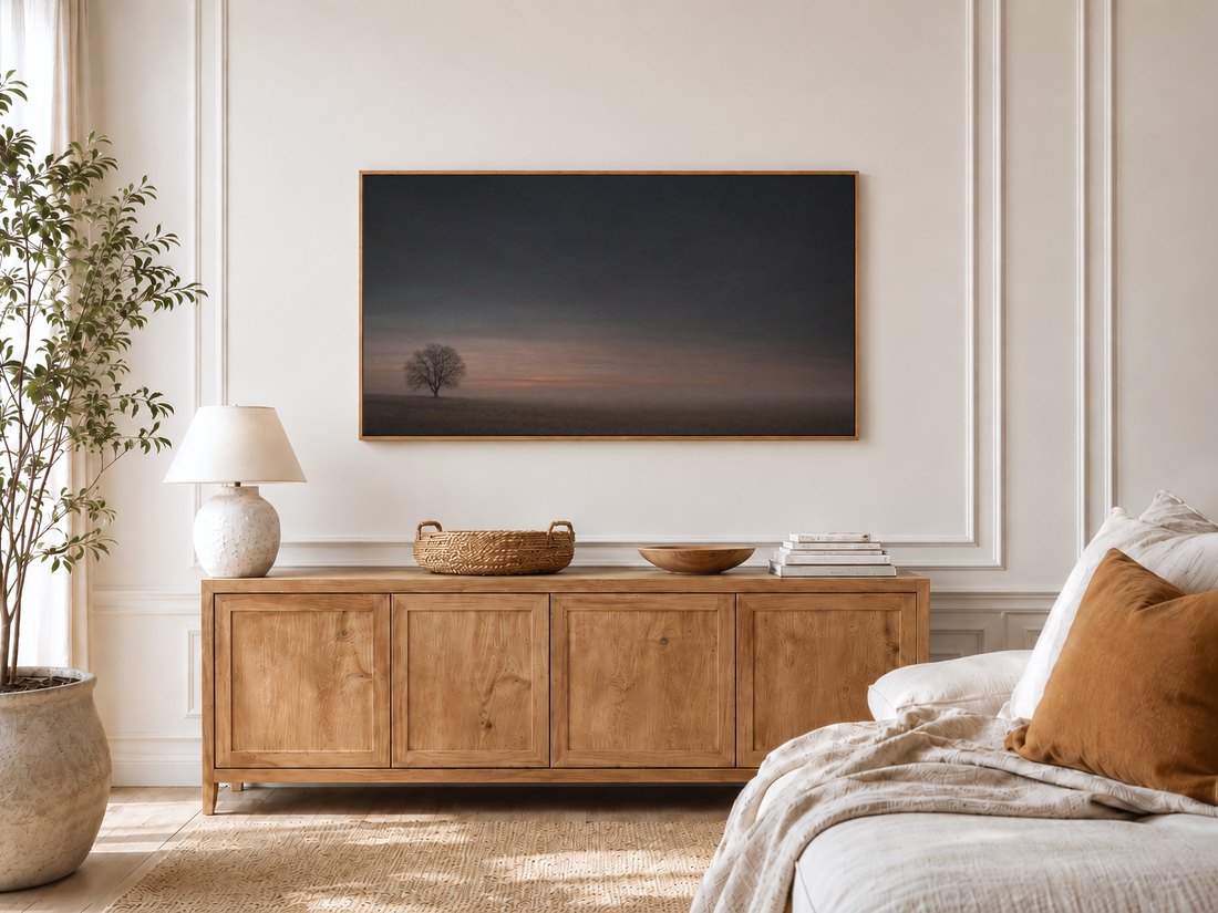 Quiet dusk field artwork on a Frame TV in a bright bedroom, above a light oak sideboard with a ceramic lamp and rattan tray, its muted grey and rust palette pairing with the linen bed and olive tree nearby.