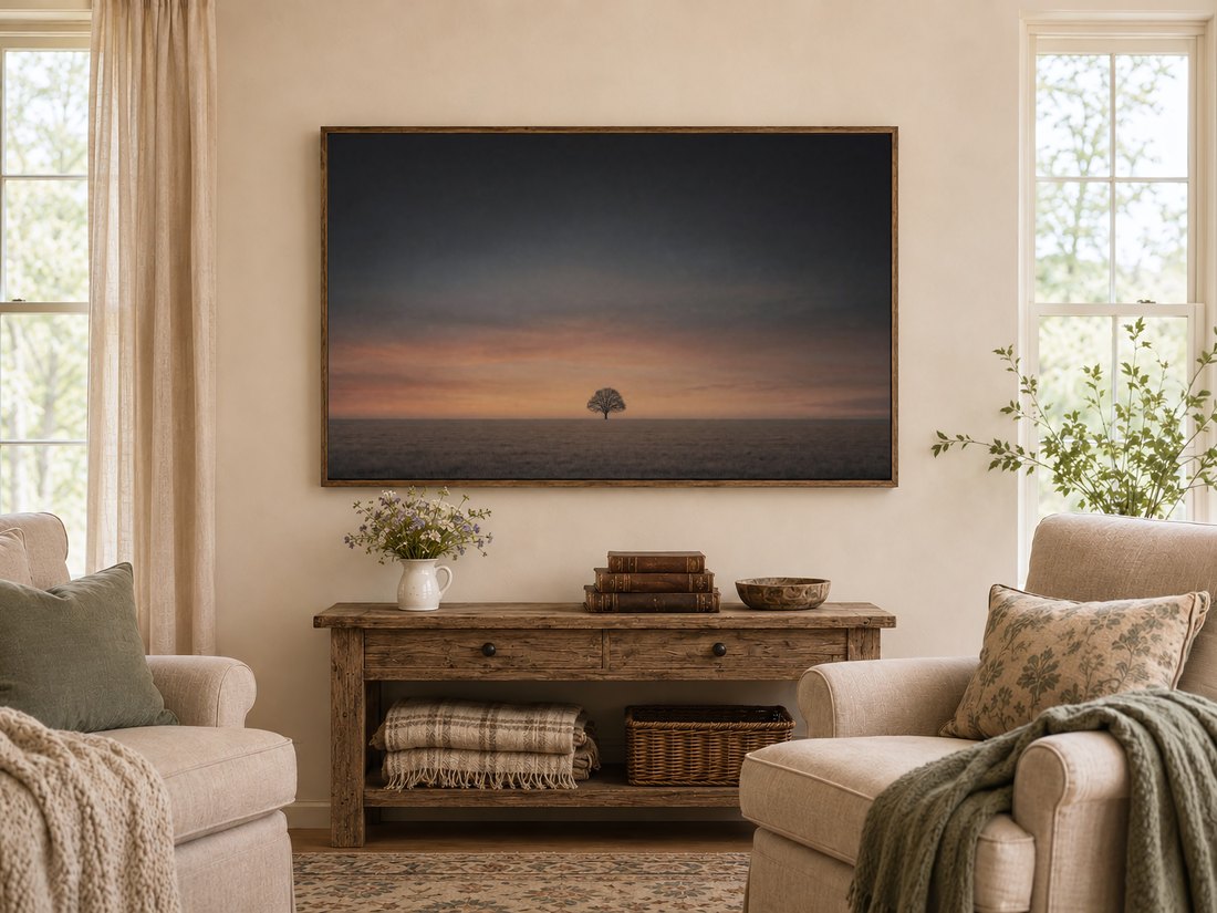 Minimalist lone tree wall art on a TV screen in a warm farmhouse living room, above a rustic console with wildflowers, leather books and a carved bowl, flanked by cream armchairs with sage cushions.