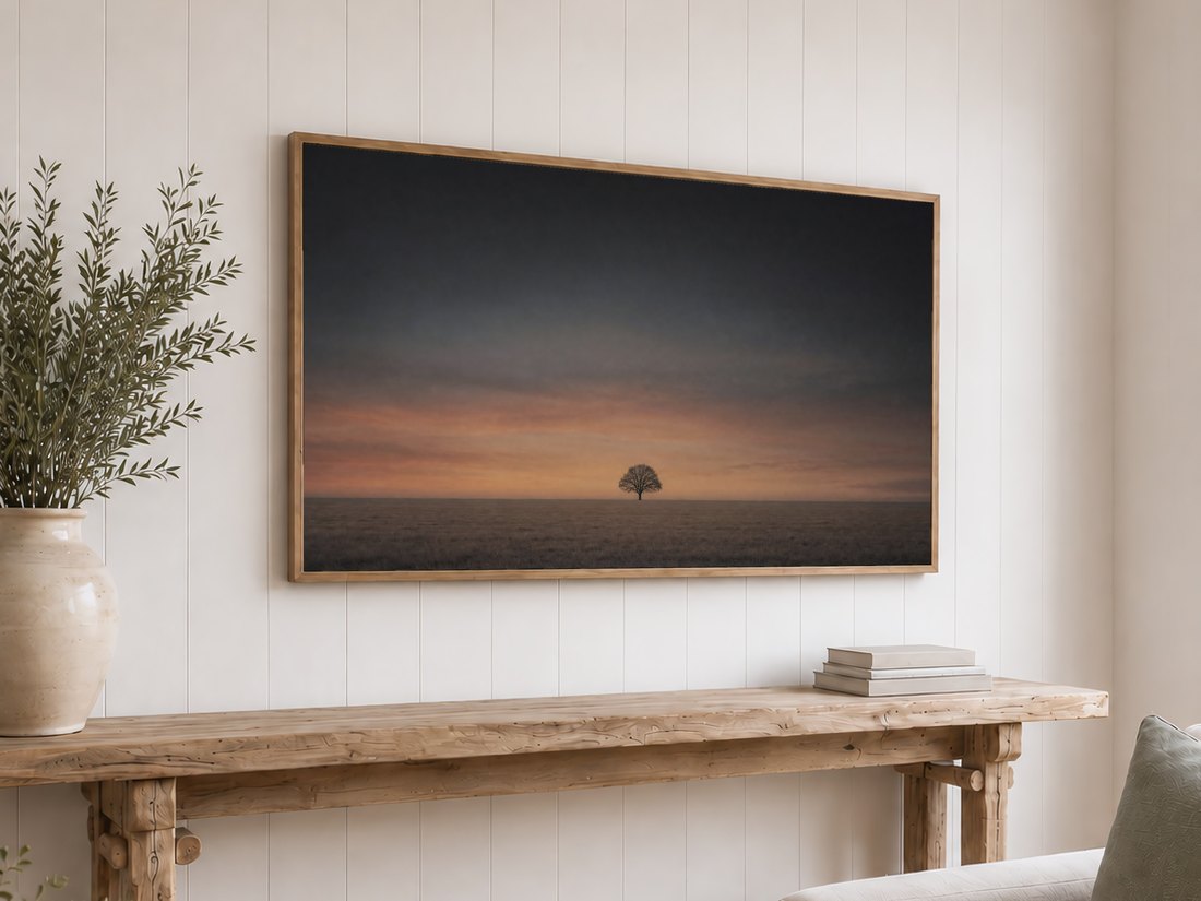 Lone tree silhouette frame TV art on a white shiplap wall in a light oak frame, above a reclaimed-wood console with olive branches and neutral books, offering a still, moody focal point.