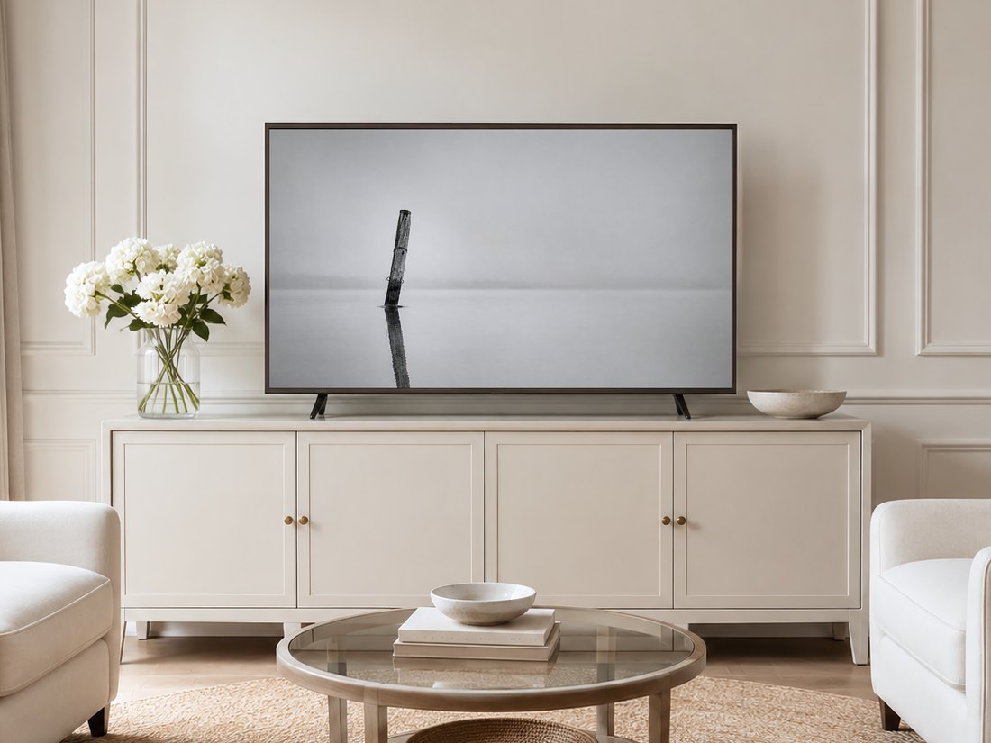 Misty black and white lake photo of a solitary mooring post on a Frame TV, set in a neutral living room with a cream panelled wall, long sideboard, white hydrangeas and pale armchairs on a jute rug.