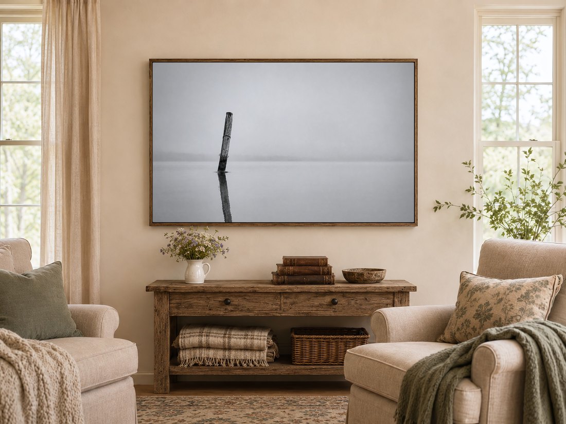 Monochrome lake art with a weathered post and still water displayed above a rustic console table in a warm farmhouse living room, near wildflowers, leather books and cream armchairs with sage cushions.