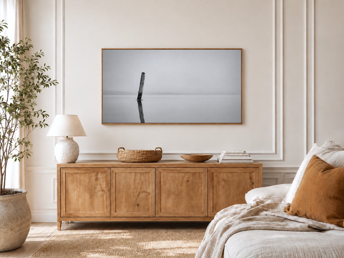 Serene black and white mooring post artwork on a Frame TV in a bright bedroom with cream panelled moulding, a light oak sideboard, ceramic lamp, potted olive tree and a linen bed with a rust cushion.