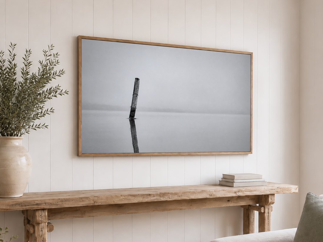 Quiet foggy lake scene in black and white shown on a light oak framed TV above a reclaimed wood console table, with olive branches and neutral books, against a white shiplap wall.