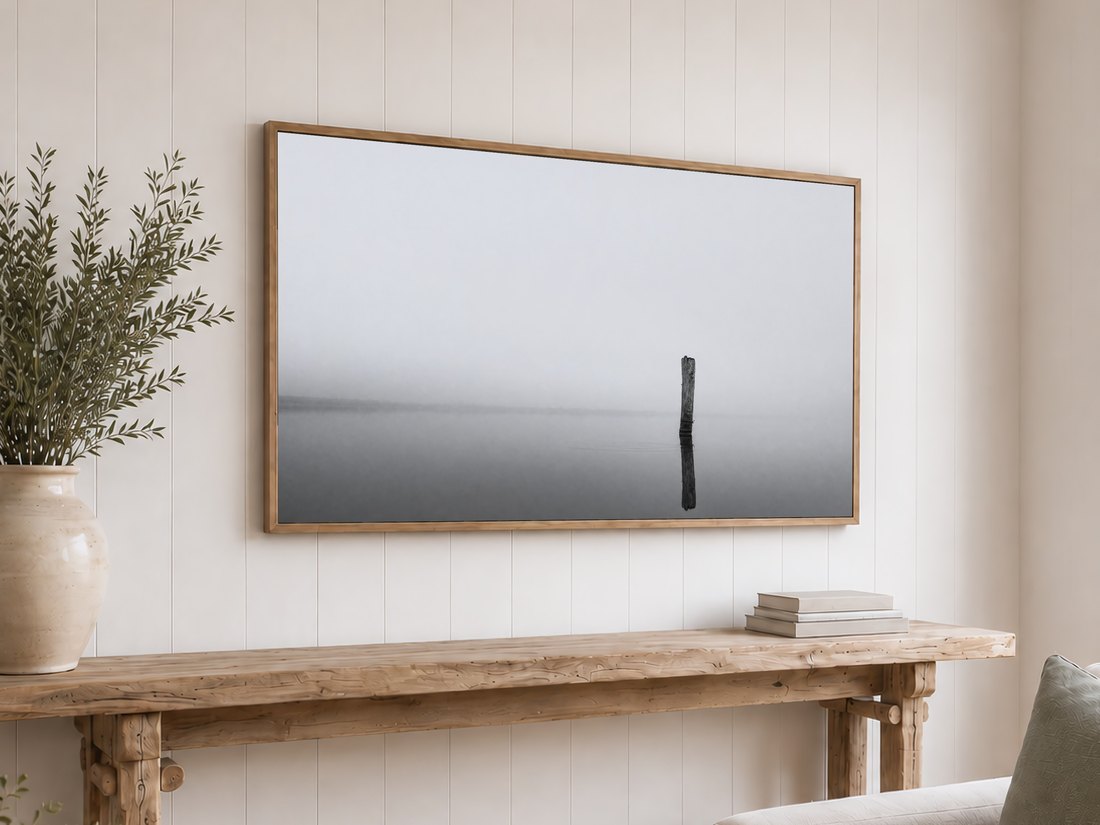 Minimalist foggy lake art displayed on a light oak framed TV above a reclaimed-wood console with olive branches and neutral books against a white shiplap wall.