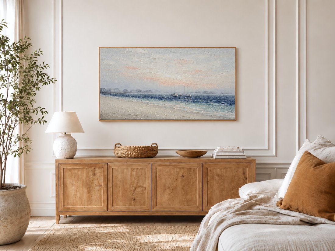 Misty coastal sailboat art on a Frame TV in a bright bedroom with cream panelled moulding, an oak sideboard, potted olive tree and linen bedding, offering a soft seaside mood to the space.