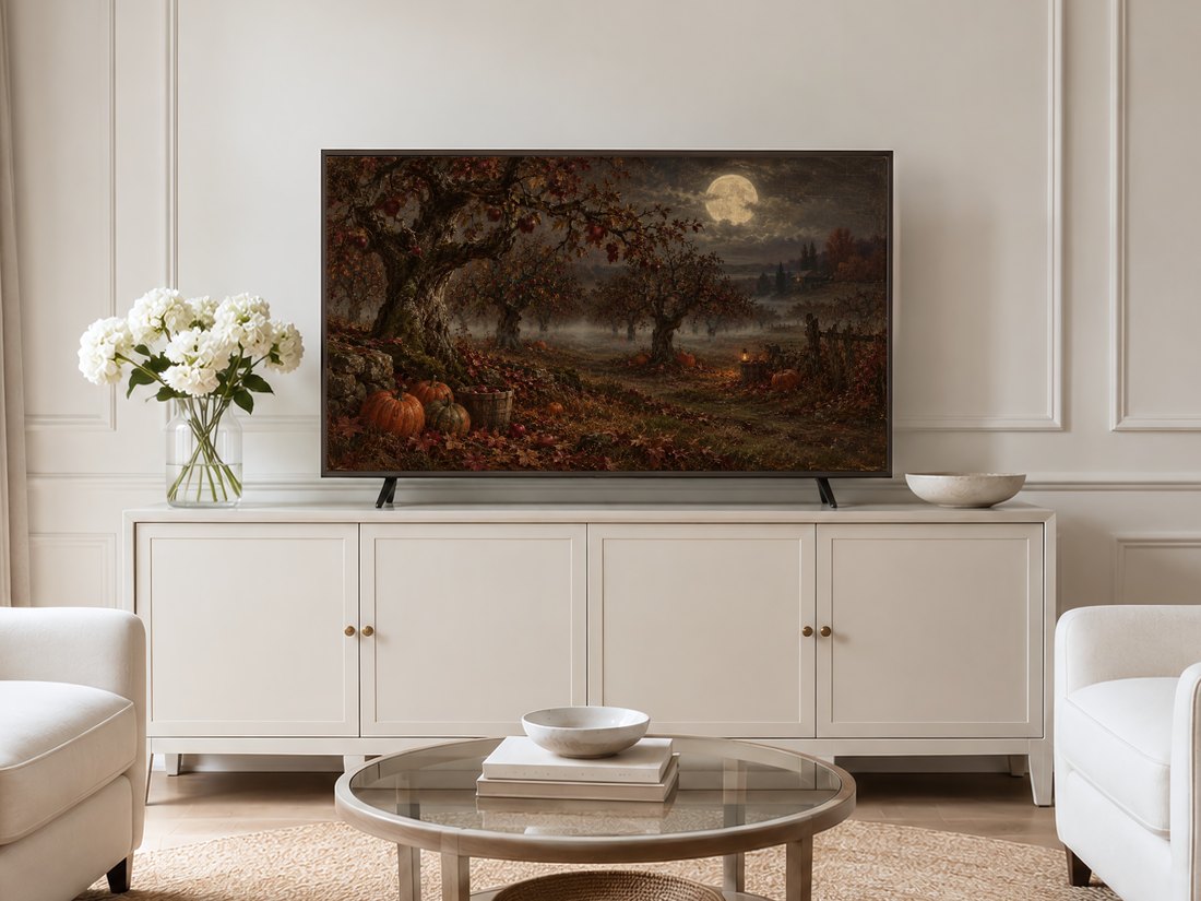 Moonlit Apple Orchard artwork shown on a Frame TV mounted on a neutral cream panelled wall above a cream sideboard with brass knobs, flanked by pale armchairs and white hydrangeas in a glass vase.
