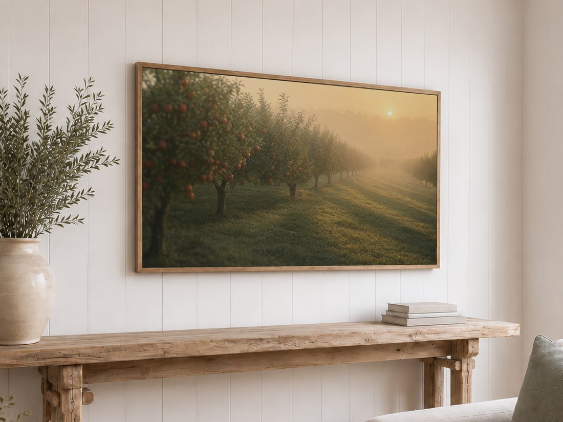 Sunrise orchard artwork on a light oak framed TV above a rustic reclaimed-wood console table with olive branches in a cream vase and a stack of neutral books against a white shiplap wall.