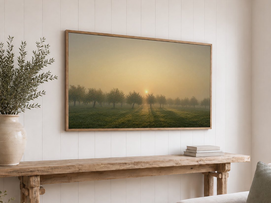 Hazy orchard sunrise shown on a light oak framed TV against a white shiplap wall, above a reclaimed-wood console with olive branches in a ceramic vase and a stack of neutral books.