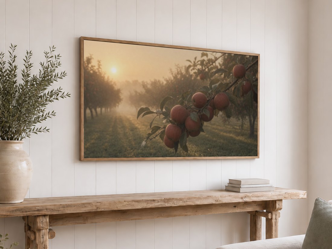 Soft apple orchard art on a light oak framed TV against a white shiplap wall, above a reclaimed-wood console with olive branches, the hazy sunrise and rust apples suiting a farmhouse living room.