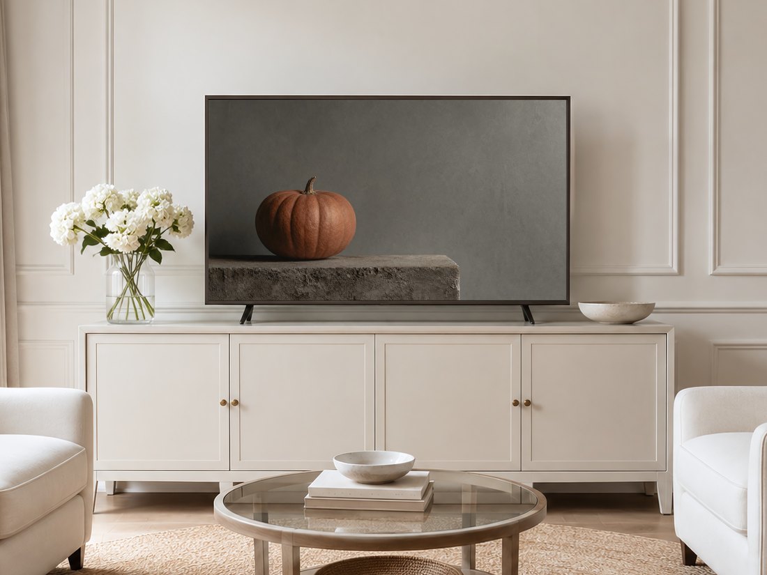 Pumpkin still life displayed on a Frame TV in a neutral living room with a cream sideboard, white hydrangeas, glass coffee table and pale armchairs on a jute rug, muted autumn accent.
