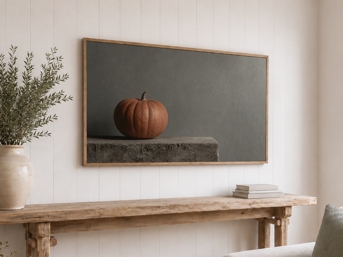 Minimalist pumpkin still life on a light oak framed TV above a reclaimed-wood console with olive branches and neutral books, set against a white shiplap wall for understated fall style.