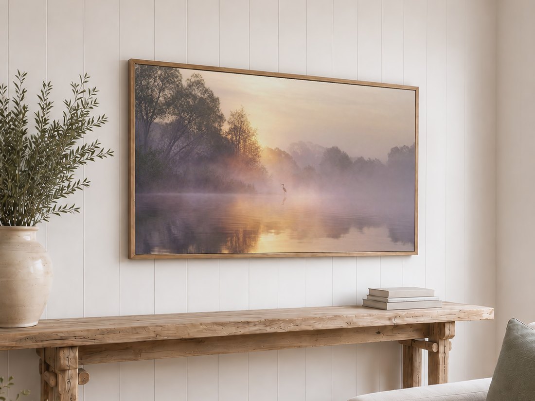Heron in morning mist shown on a light oak framed Frame TV above a reclaimed-wood console with olive branches and neutral books, the hazy lake tones pairing well with a white shiplap wall.