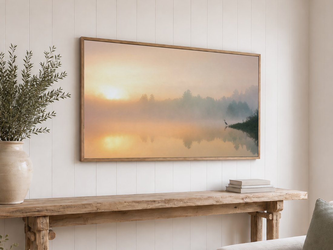 Misty sunrise lake art with a heron displayed on a light oak framed TV above a reclaimed-wood console, white shiplap wall, olive branches in a ceramic vase and a stack of neutral books.