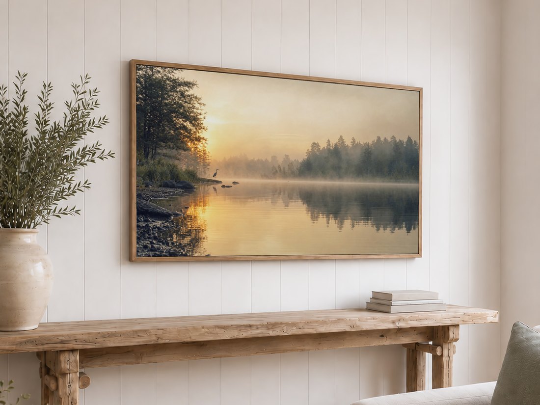 Amber-toned misty lake scene with a heron shown on a light oak framed TV above a reclaimed-wood console table with olive branches in a cream vase and a stack of neutral books, against a white shiplap wall.