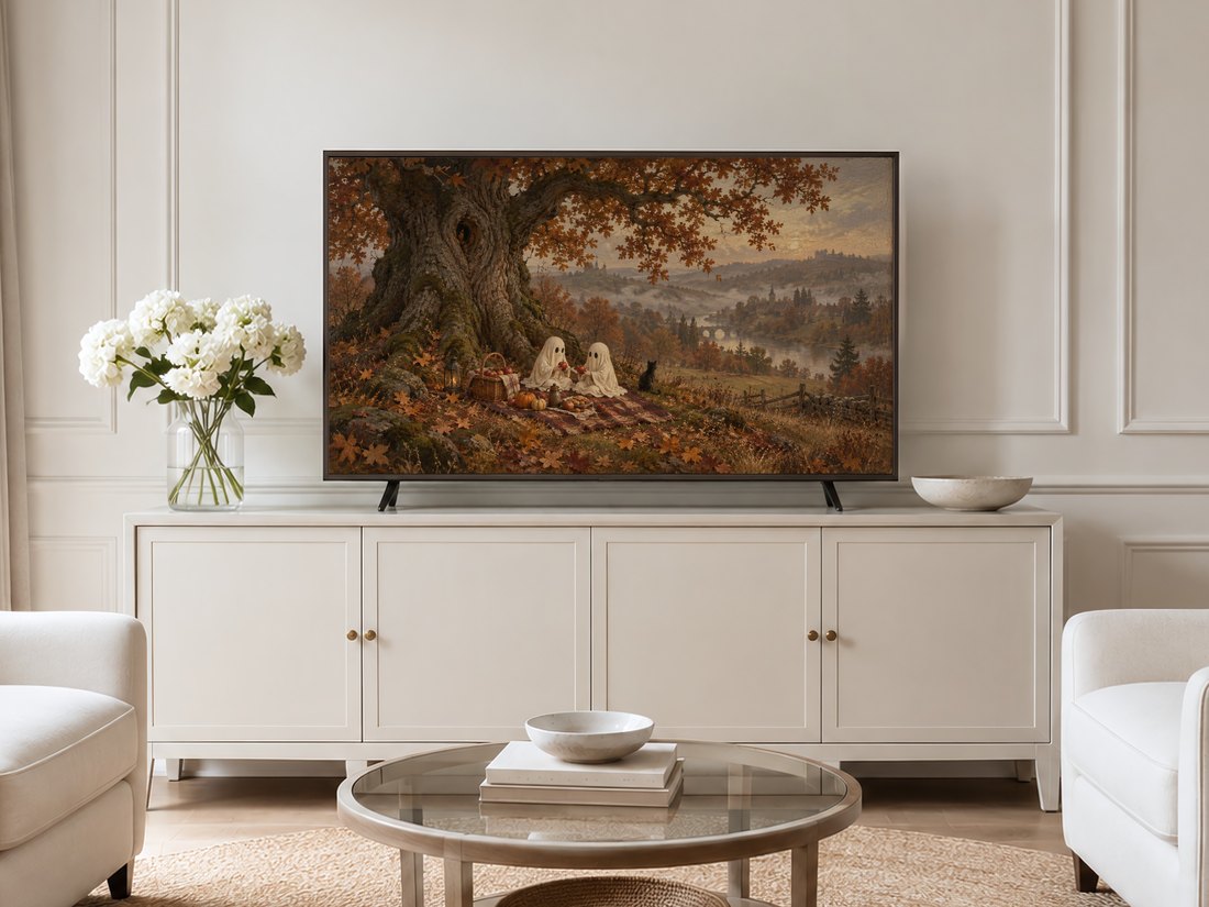 Autumn ghost picnic landscape art displayed in a thin gold frame on a neutral cream panelled living room wall above a cream sideboard, flanked by pale armchairs and white hydrangea flowers.