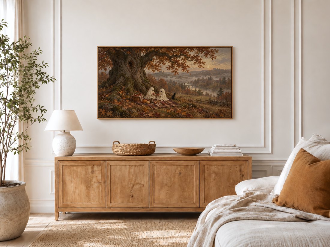 Autumn landscape with ghostly picnic scene displayed in a thin frame above a light oak sideboard in a bright bedroom with cream moulding, textured ceramic lamp, potted olive tree and linen bed.