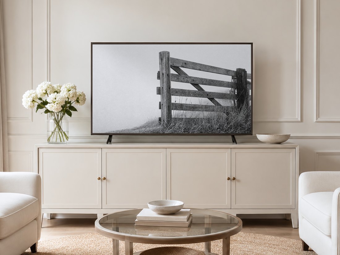 Black and white weathered fence photograph displayed on a Frame TV in a neutral living room with cream panelled walls, cream sideboard, white hydrangeas in glass vase, round glass coffee table and pale armchairs on jute rug.