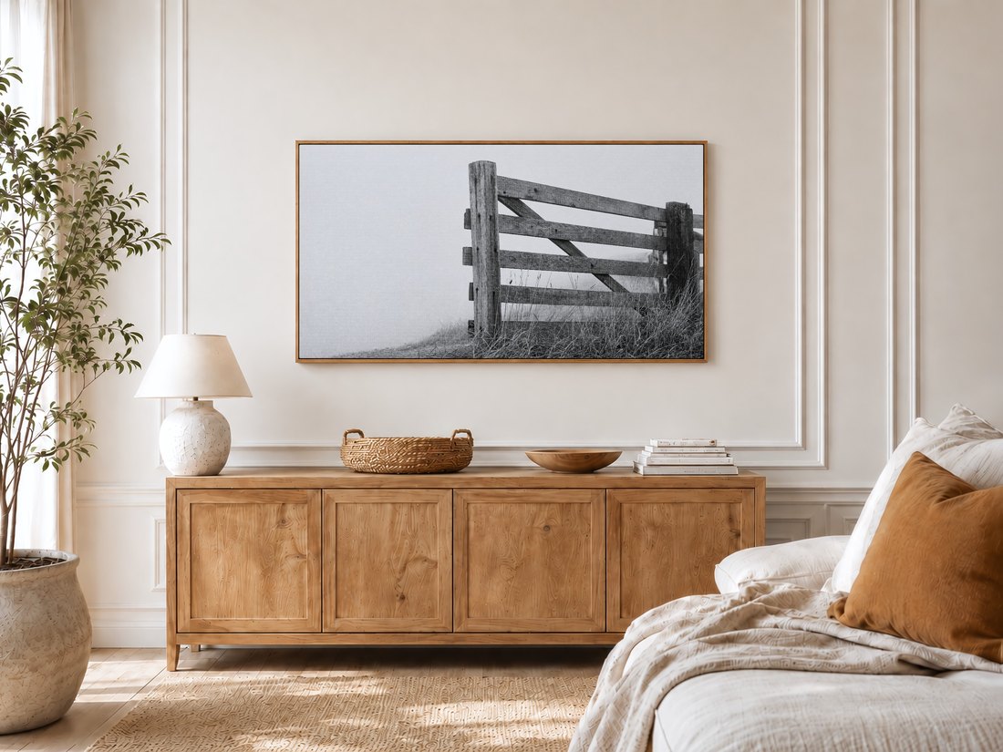 Black and white fence photograph on Frame TV in a bright bedroom with cream panelled moulding, light oak sideboard with ceramic lamp and rattan tray, potted olive tree by the window and linen bed with rust cushion.