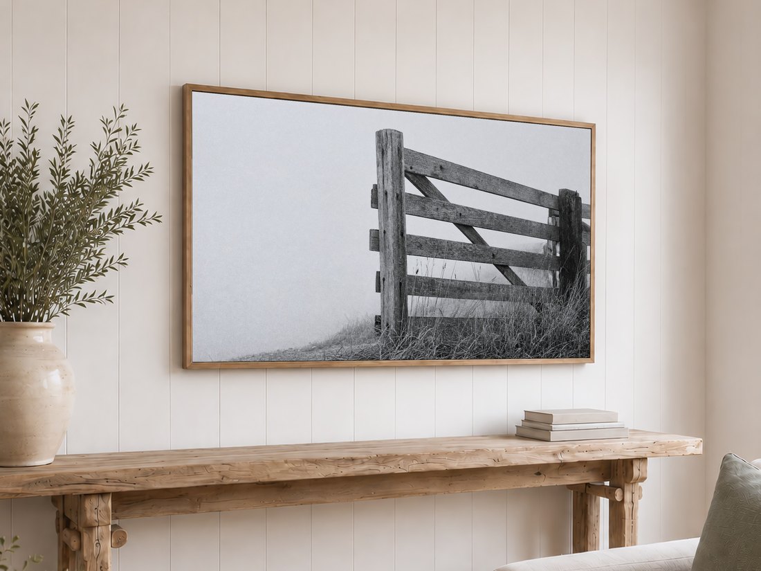 Weathered fence in fog displayed on Frame TV above a rustic reclaimed-wood console table on a white shiplap wall, with light oak frame, olive branches in cream ceramic vase and stack of neutral books nearby.