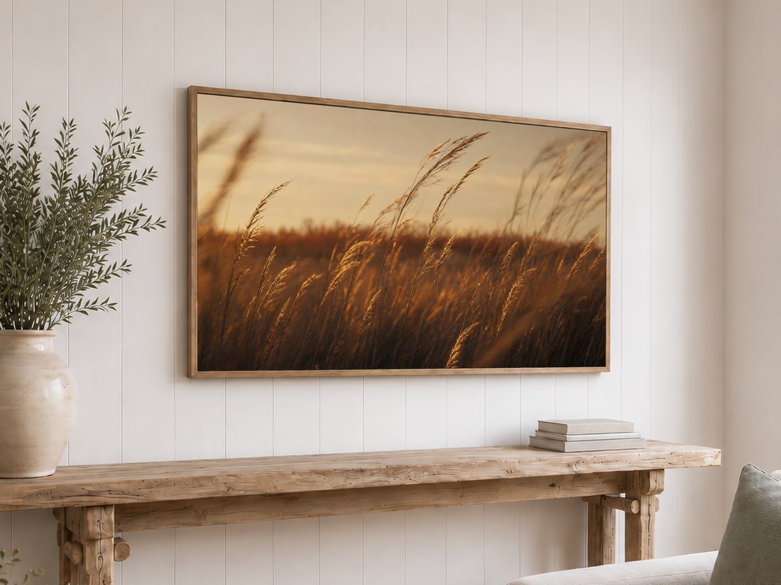 Golden prairie grass artwork on an oak framed TV above a reclaimed-wood console table with olive branches in a cream vase and neutral books, set against a white shiplap wall for a warm, natural look.