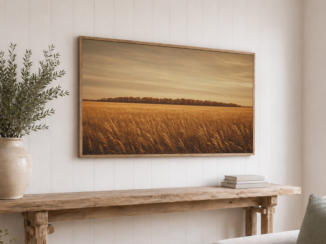 Warm golden field artwork with a hazy sunset sky shown on a light oak framed TV above a reclaimed-wood console with olive branches and neutral books against a white shiplap wall.